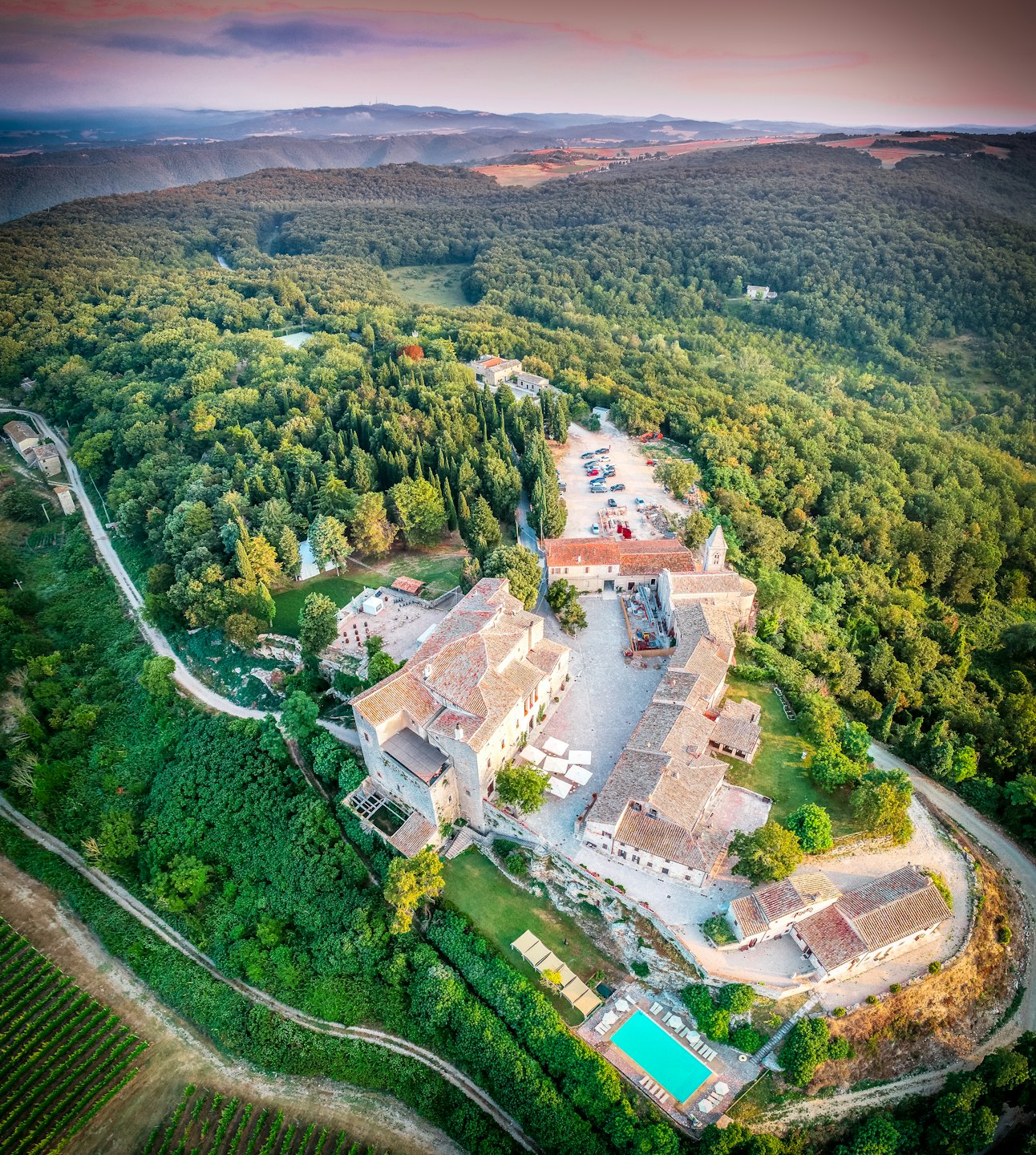 Antica tenuta in pietra con tetti in cotto e piscina blu circondata da boschi e vigneti.